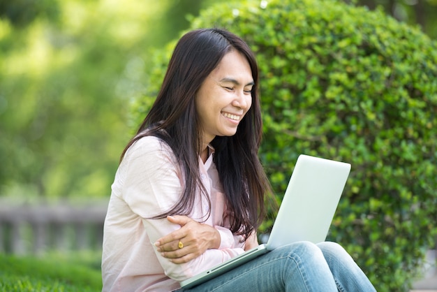 Junge asiatische schöne Frau mit lächelndem Gesicht, das draußen in einem öffentlichen Park arbeitet. Arbeiten am Laptop im Freien. Beschnittenes Bild der Frau, die am Laptop arbeitet, während sie in einem Park sitzt.