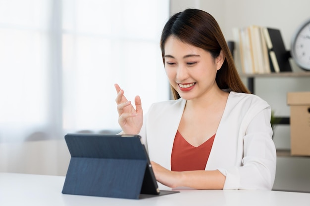 Junge asiatische schöne Geschäftsfrau, die mit dem Tablet arbeitet, das zu Hause sitzt. Lächelnde, charmante, glückliche junge Frau, die zu Hause eine Hausaufgabenkonferenz mit dem Team macht.