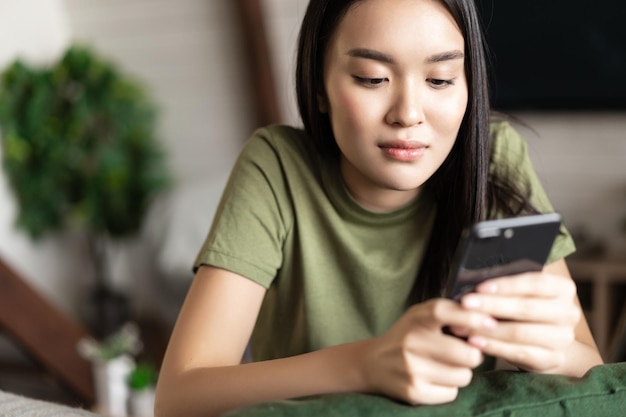 Junge asiatische Studentin, die auf den Handybildschirm schaut, der zu Hause im Wohnzimmer ruht?