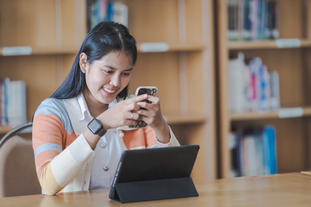 Junge asiatische Studentin in Studentenuniform, die online studiert, ein Buch liest, ein digitales Tablet oder einen Laptop in der Universitätsbibliothek verwendet, während das Klassenzimmer während der COVID-19-Pandemie eingeschränkt ist