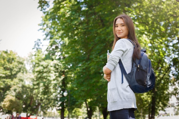 Junge asiatische studentin, mit büchern und rucksack. nach dem unterricht im park bleiben und lächeln. nachdenken über den kommenden tag. lifestyle- und bildungskonzept