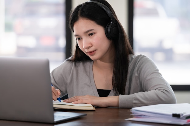 Junge asiatische Studentin trägt drahtlose Kopfhörer, schreibt auf dem Notebook, um die Sprache online zu lernen und dem Dozenten zuzuhören, Webinar per Videoanruf, E-Learning zu Hause, Fernunterricht