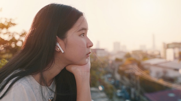 Junge asiatische tausendjährige gen z Frau, die am Dach steht, sich gelangweilt fühlt und Stadtbild betrachtet, während Musik hört