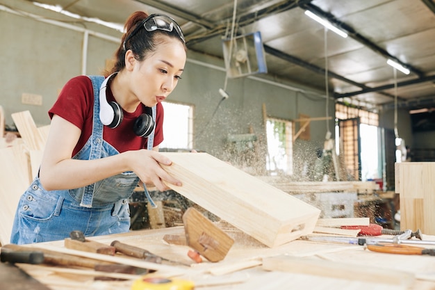 Junge asiatische Tischlerin bläst Holzstaub aus der Schublade, die sie in der Tischlerei hergestellt hat
