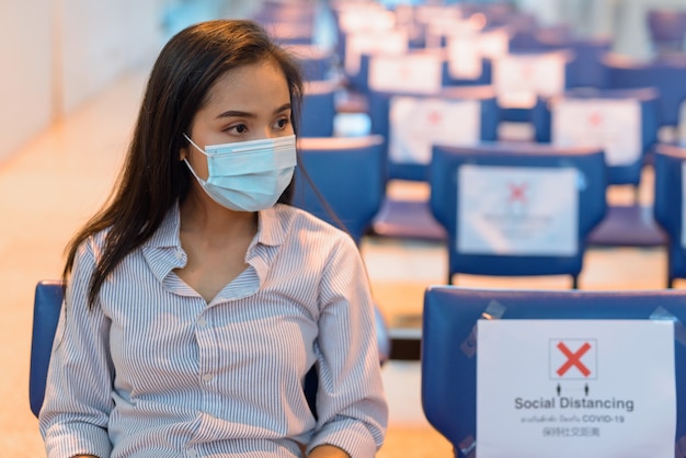Junge asiatische touristenfrau, die maske trägt und mit abstand am flughafen sitzt