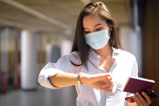 Junge asiatische Touristenfrau mit Maskenprüfzeit beim Halten des Passes am Flughafen