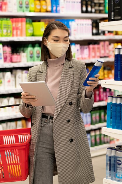 Junge asiatische Verbraucherin mit rotem Korb und Tablette, die an einem großen Regal mit Haarpflegeprodukten steht und ihre Eigenschaften durchschaut