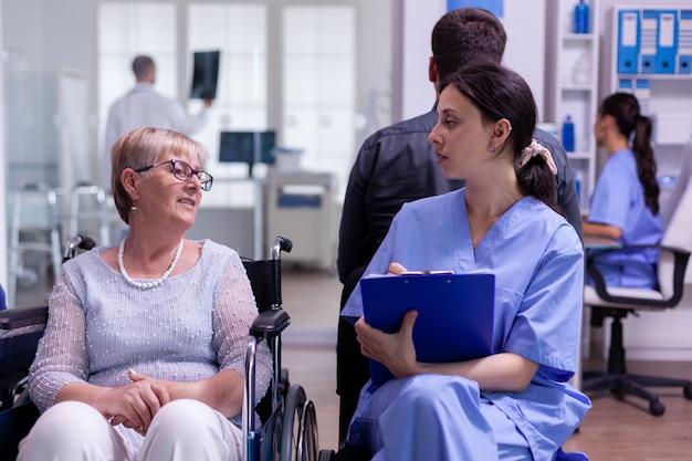 Junge Assistentin, die das Registrierungsformular einer behinderten älteren Patientin überprüft, die im Rollstuhl im Wartebereich des Krankenhauses sitzt sitting