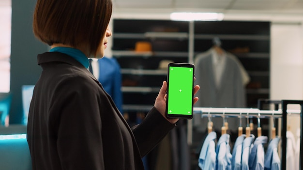 Junge Assistentin verwendet leere Greenscreen-Vorlage und zeigt Chroma-Key-Display in trendigem Bekleidungsgeschäft. Asiatischer Arbeiter nutzt isolierten Mockup-Copyspace auf modernem Smartphone, Einkaufszentrum.