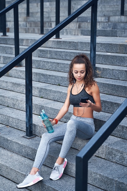 Foto junge athletische frau in sportkleidung, die auf der treppe des stadtstadions mit telefon in den händen sitzt