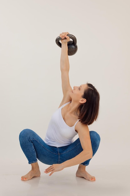 Junge athletische Frau sitzt und hebt den weißen Hintergrund der Kettlebell hoch und hält sich durch Krafttraining fit