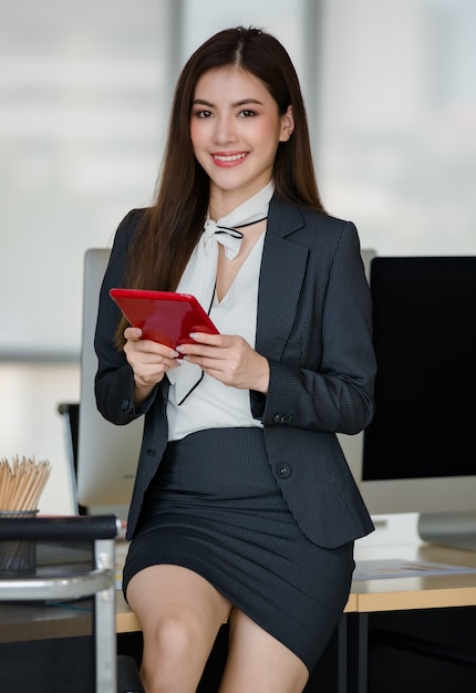 Junge attraktive Asiatin im schwarzen Anzug, die an roter Tablette in modern aussehendem Büro mit verschwommenem Fensterhintergrund arbeitet. Konzept für modernen Bürolebensstil.