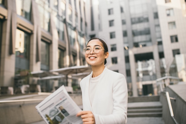 Junge attraktive asiatische Bankierin oder Buchhalterin in Gläsern liest Zeitung außerhalb eines modernen Bürozentrums