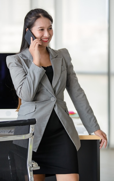 Junge attraktive asiatische Frau im grauen Anzug, die am Handy im modernen schauenden Büro mit unscharfem Fensterhintergrund spricht. Konzept für modernen Bürolebensstil.