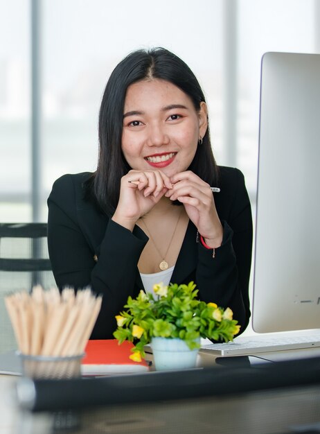 Junge attraktive asiatische Frau im schwarzen Anzug, die mit Zuversicht in modern aussehendem Büro mit verschwommenem Fensterhintergrund sitzt. Konzept für modernen Bürolebensstil.