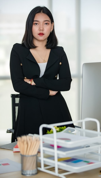 Junge attraktive asiatische Frau im schwarzen Anzug stehend Arm gekreuzt mit Vertrauen in modern aussehendes Büro. Konzept für modernen Bürolebensstil.