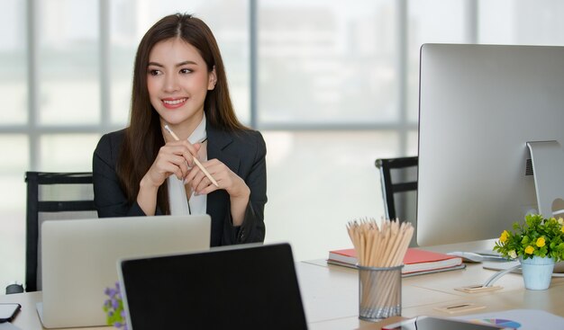 Junge attraktive asiatische Frau im schwarzen Geschäft, das hinter Tisch mit Computer und Laptop im modernen schauenden Büro mit undeutlichem Fensterhintergrund sitzt. Konzept für modernen Bürolebensstil.