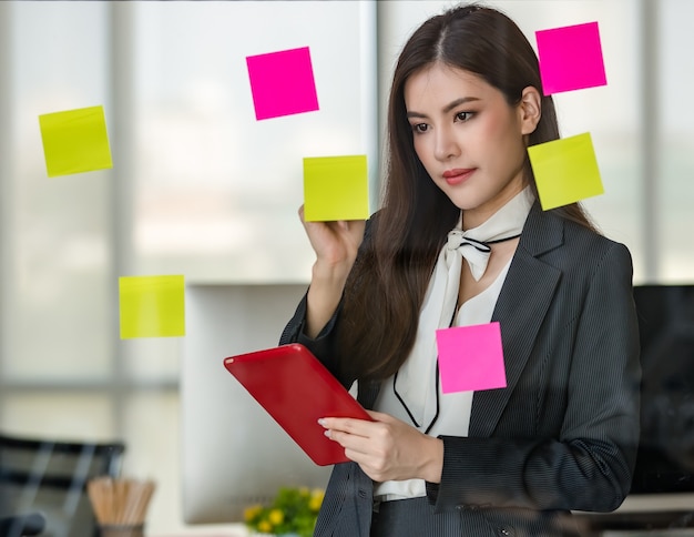 Junge attraktive asiatische Frau im schwarzen Geschäftsschreiben auf Post-It auf Glasplatte eine Hand, die Tablette in modernem schauendem Büro mit unscharfem Fensterhintergrund hält. Konzept für modernen Bürolebensstil.