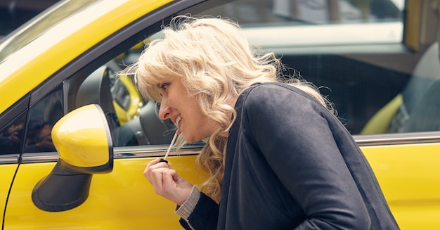 Foto junge attraktive blonde frau malt ihre lippen, die den seitenspiegel eines gelben autos betrachten