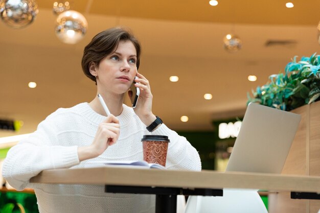 Junge attraktive brünette Frau, die in einem Einkaufszentrum an einem Tisch sitzt und an einem Computer arbeitet