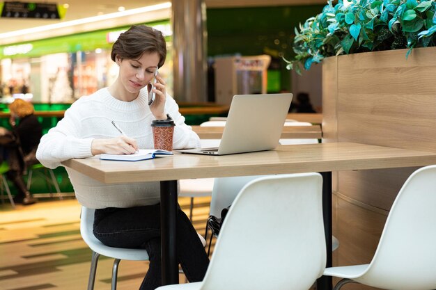 Junge attraktive brünette Frau sitzt in einem Einkaufszentrum an einem Tisch und arbeitet an einem Computer-Laptop, benutzt ein Handy und schreibt Informationen auf ein Notizbuch. Freiberufliches und Geschäftskonzept