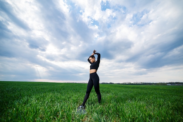 Junge attraktive Brunettefrau, die in der Natur steht und schwarze Gamaschen und ein schwarzes Oberteil trägt. Sommertag, grünes Gras. Athletischer Körper und gesunder Lebensstil.