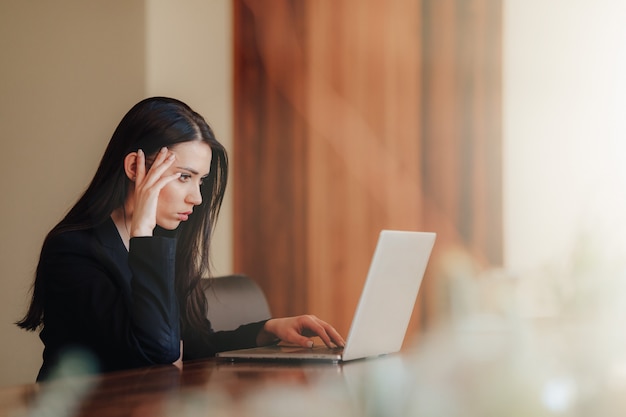 Junge attraktive emotionale Frau in der Geschäftsart Kleidung, die am Schreibtisch mit einem Laptop sitzt