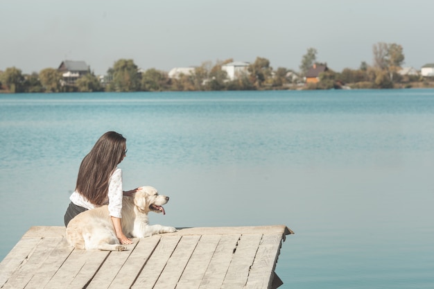 Junge attraktive Frau, die am Pier mit ihrem Hund sitzt. Beste Freunde im Freien