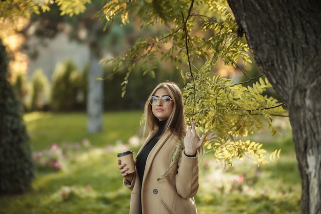 Junge attraktive Frau gekleidet im stilvollen Mantel, Rollkragenpullover, der im Herbstpark aufwirft und Tasse Kaffee hält