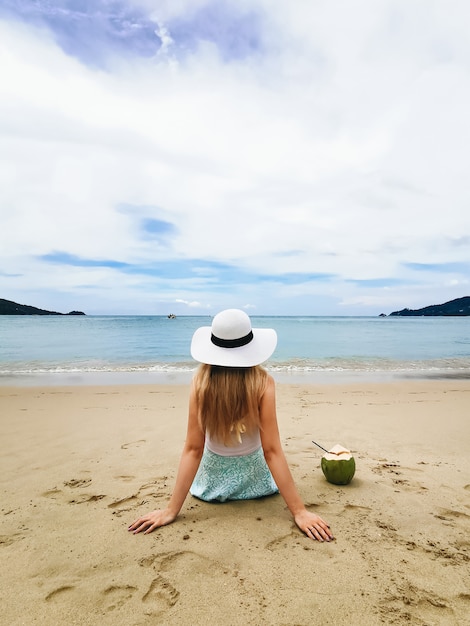 Junge attraktive Frau in einem Hut am Ufer des blauen Meeres, das zurück mit einer Kokosnuss, Thailand sitzt