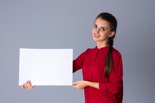 Junge attraktive Frau in roter Bluse mit Zopf mit weißem Blatt Papier auf grauem Hintergrund im Studio