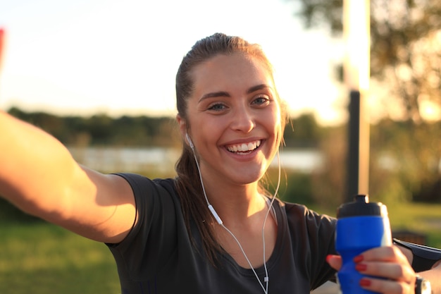 Junge attraktive Frau in Sportkleidung, die auf das Telefon schaut und lächelt, während sie Selfie macht, während des Trainings im Freien