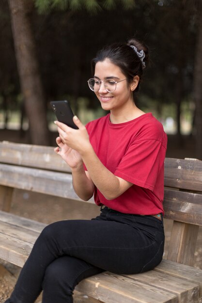 Junge attraktive Frau mit Brille, die in einer Bank sitzt und ihr Telefon in einem Park mit einem roten Hemd benutzt. Sevilla, Spanien