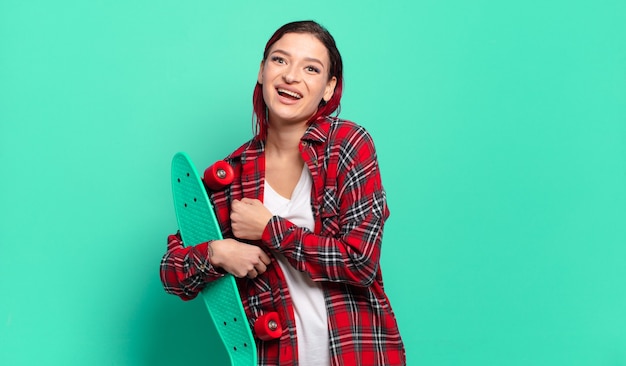 Junge attraktive Frau mit roten Haaren, die sich glücklich, positiv und erfolgreich fühlt, motiviert oder gute Ergebnisse feiert und ein Skateboard hält