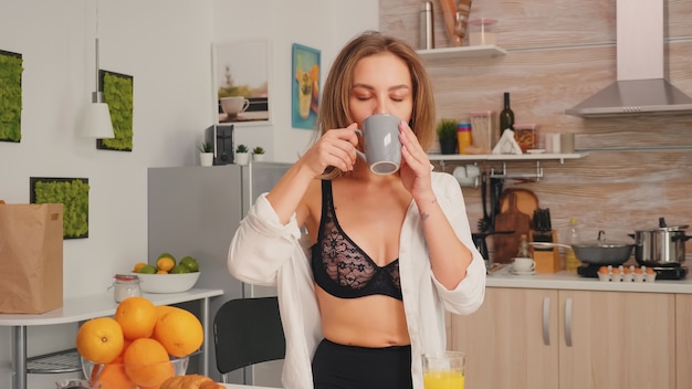 Junge attraktive Frau mit Tätowierungen in verführerischer Unterwäsche, die eine Tasse Tee in der Küche lächelnd hält. Sexy blonde Dame in der Wäsche, die Kaffee während des Frühstücks trinkt, das den Morgen genießt.