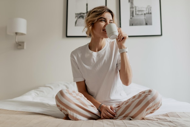 Junge attraktive glückliche Frau mit hellem Haar sitzt morgens im Bett und trinkt Kaffee nach dem Aufwachen am Morgen Glücklicher sonniger Morgen zu Hause