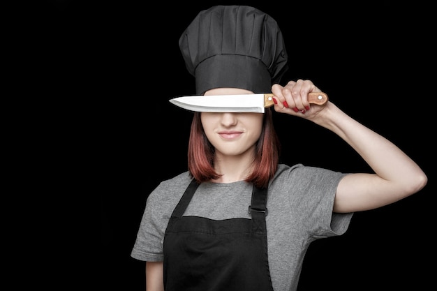 Junge attraktive Köchin in schwarzer Uniform hält Messer vor Gesicht auf schwarzem Hintergrund
