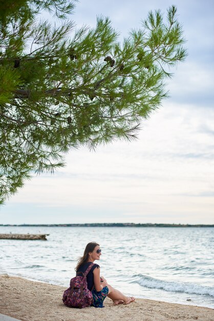 Junge attraktive lächelnde Frau mit buntem Rucksack, der allein auf sandigem Ozeanstrand auf hellblauem klarem Wasser und Himmel sitzt