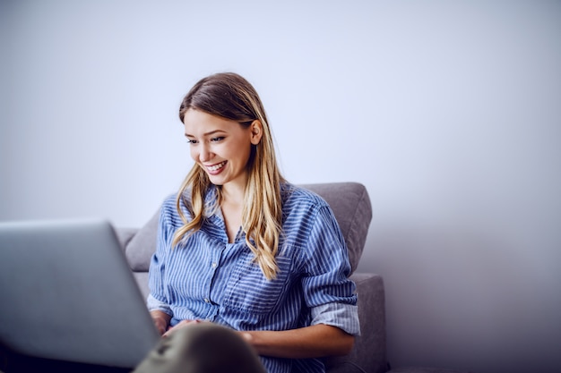 Junge attraktive lächelnde kaukasische Brünette, die auf Sofa im Wohnzimmer sitzt und Laptop für freiberuflichen Job verwendet.