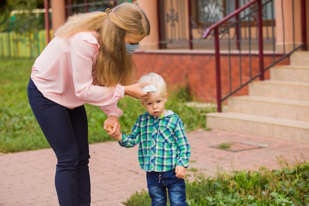 Junge attraktive Lehrerin, die die Temperatur des kleinen Jungen mit einem berührungslosen Termometer misst Neue Regeln zum Schutz von Kindern im Kindergarten Neues Normalkonzept während einer Pandemie