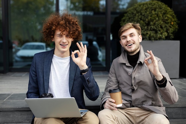 Junge attraktive Männer mit Laptop, die im Freien auf der Straße eine Ok-Geste zeigen Konzept des Online-Arbeitsfreiberuflers