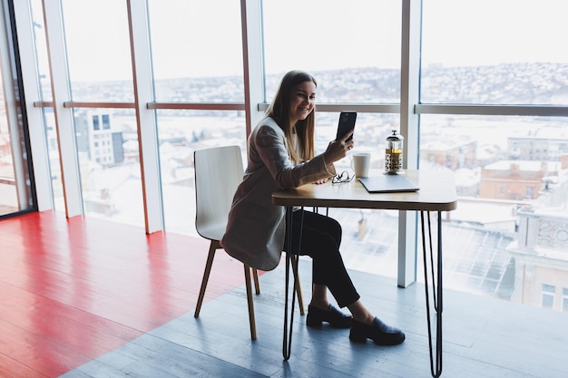 Junge attraktive Managerin spricht auf einem Mobiltelefon und lächelt, während sie in ihrer Freizeit allein in der Nähe eines großen Fensters in einem Café sitzt und an einem Computer arbeitet Glückliche Frau ruht sich in einem Café aus