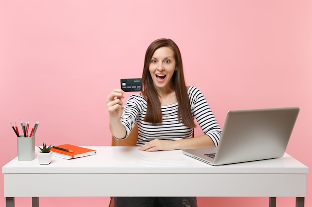 Junge aufgeregte Frau, die Kreditkarte hält, während sie am weißen Schreibtisch mit zeitgenössischem PC-Laptop sitzt, einzeln auf pastellrosa Hintergrund. Erfolgsgeschäftskarrierekonzept. Kopieren Sie Platz für Werbung.