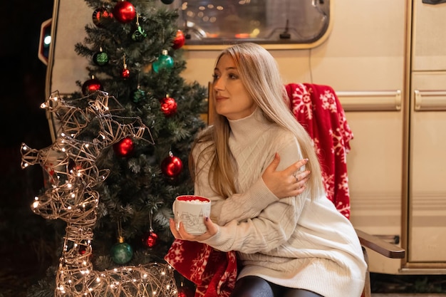 Junge aufrichtige Frau in gestricktem, kuscheligem Pullover, die in den Händen eine Tasse Kakaogetränk hält und sich selbst umarmt, entspannt auf einem Stuhl sitzend, der Outdoor-Genuss macht, frohes neues Jahr und frohe Weihnachtsfeiertage