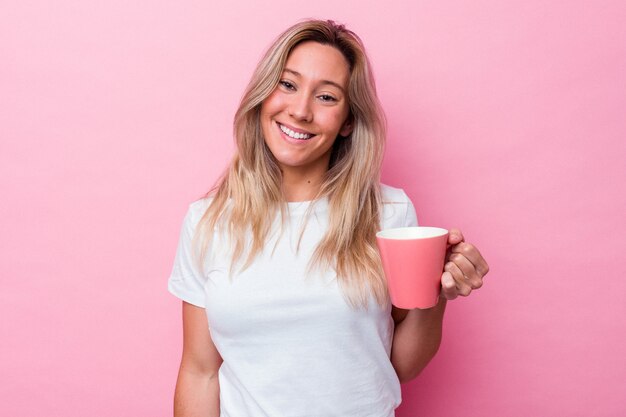 Junge australische Frau, die einen rosafarbenen Becher lokalisiert auf rosa Hintergrund hält glücklich, lächelnd und fröhlich.