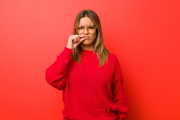 Junge authentische charismatische wirkliche Leutefrau gegen eine Wand mit den Fingern auf den Lippen, die ein Geheimnis halten.