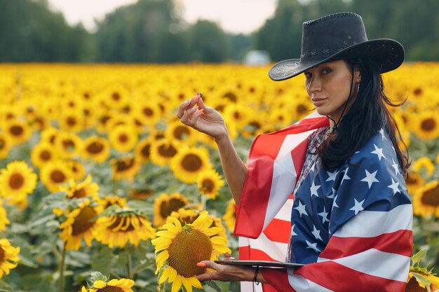 Junge Bäuerin mit amerikanischer Flagge und Tablet-PC im Sonnenblumenfeld