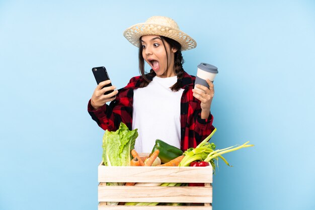 Junge Bäuerin mit frischem Gemüse in einem Holzkorb mit Kaffee zum Mitnehmen und einem Handy and