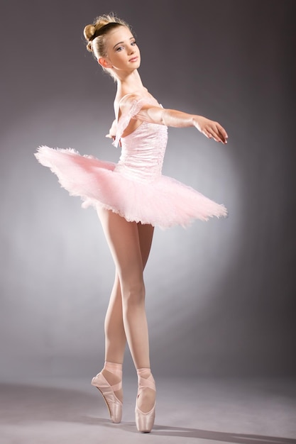 Junge Ballerina in weißem Tutu tanzt auf Pointe mit Armen über dem Kopf im Studio vor dunklem Hintergrund