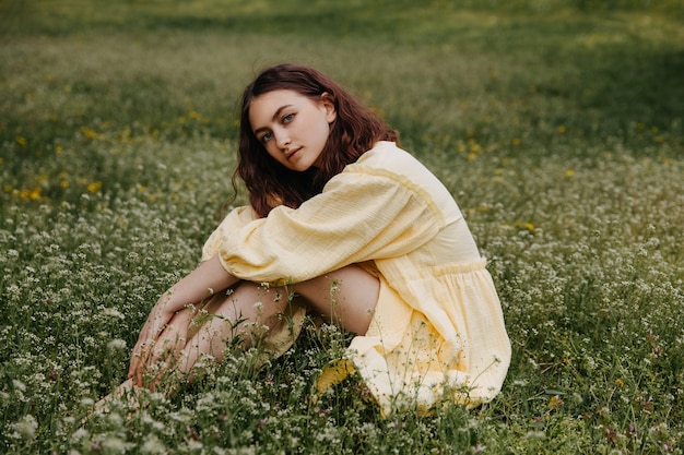 Junge barfüßige Frau, die ein gelbes Kleid trägt und auf einem Feld mit grünem Gras sitzt sitting